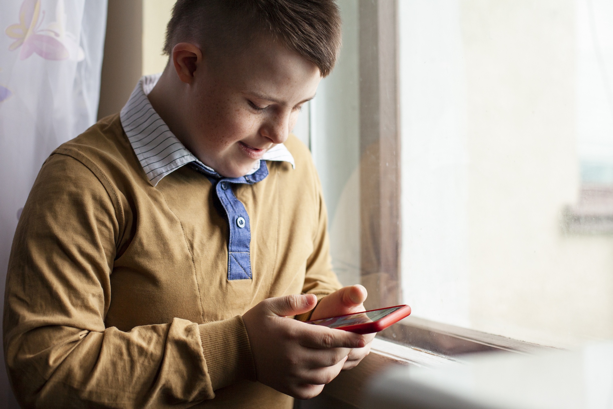 A young boy with Down syndrome smiling while using a smartphone by a window